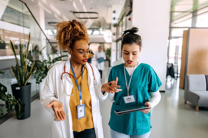 A woman wearing a white lab coat looks at a tablet with a woman in hospital scrubs.