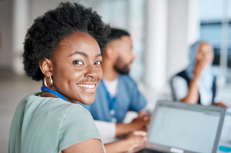 African American woman wearing scrubs in classroom with laptop, looking over shoulder at camera and smiling