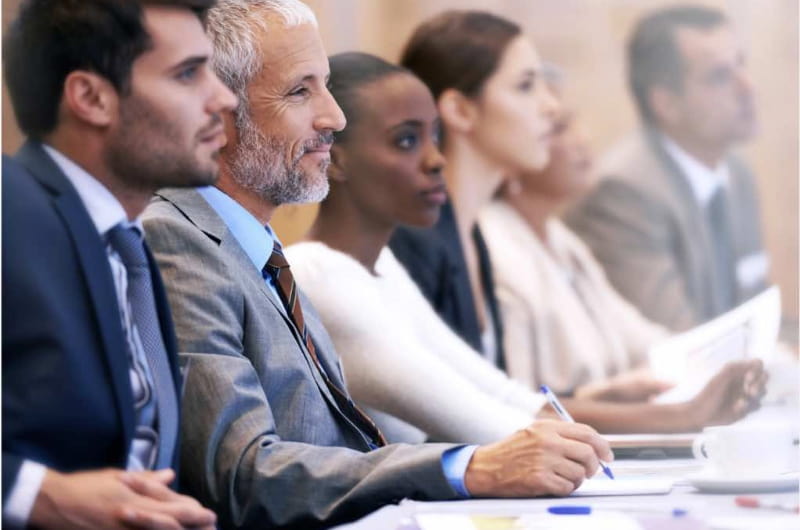 A diverse group of professionals sit at a table taking notes