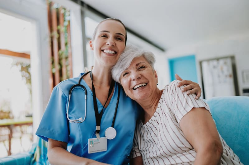 Smiling, young female healthcare worker in scrubs, sitting next to smiling elderly woman, arm around shoulders