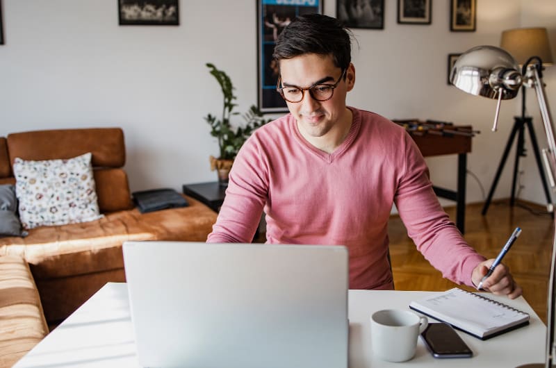 Man wearing glasses in casual clothing, writing notes while reading on a laptop