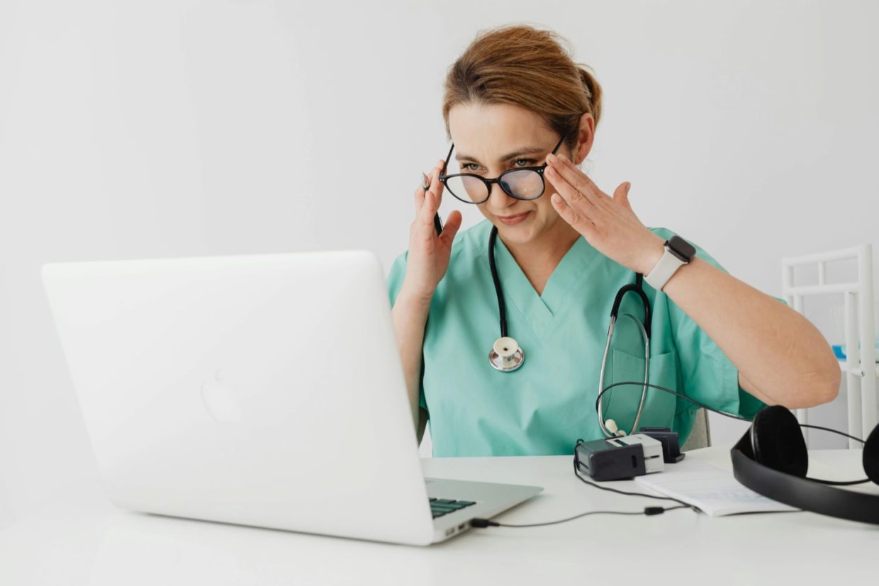 Woman in scrubs with glasses in front of laptop computer.